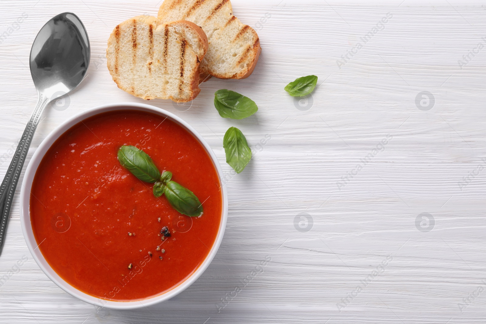 Photo of Delicious tomato cream soup in bowl served with pieces of grilled bread on white wooden table, flat lay. Space for text