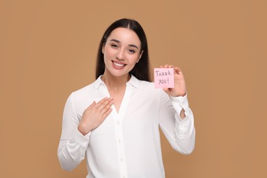 Photo of Happy woman holding paper note with phrase Thank You on brown background