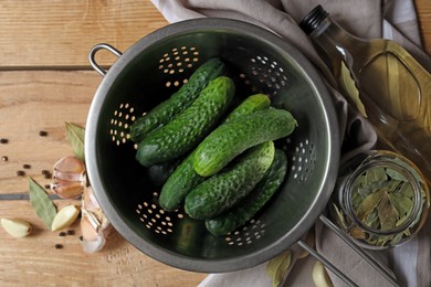Photo of Fresh cucumbers in colander and other ingredients prepared for canning on wooden table, flat lay