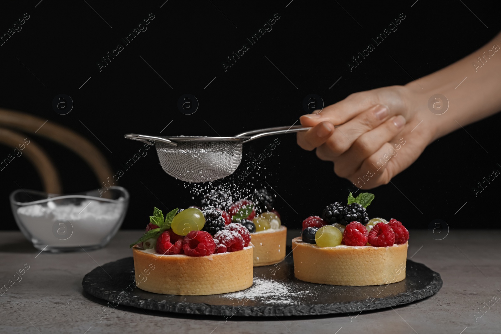Photo of Woman sprinkling delicious tartlets with powdered sugar at grey table, closeup
