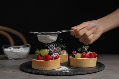 Woman sprinkling delicious tartlets with powdered sugar at grey table, closeup