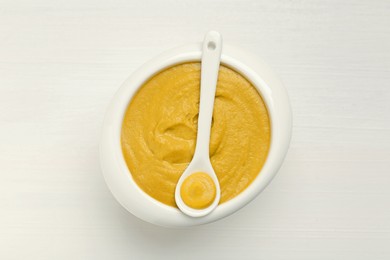 Photo of Spoon and bowl of tasty mustard sauce on white wooden table, top view