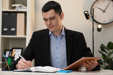 Photo of Man taking notes while using tablet at table in office