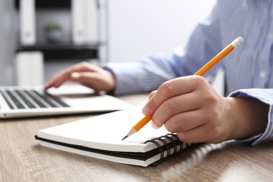 Woman with notebook working on laptop at wooden table in office, closeup