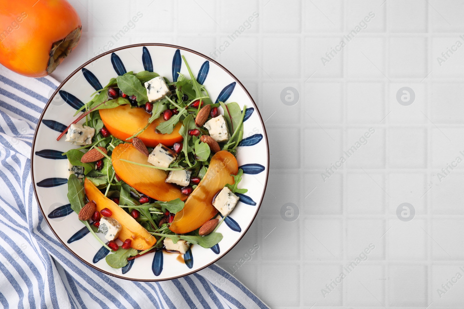 Photo of Tasty salad with persimmon, blue cheese, pomegranate and almonds served on white tiled table, top view. Space for text