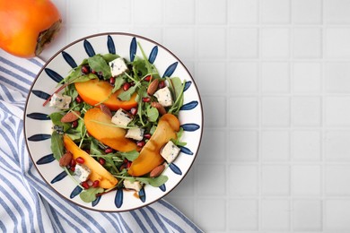 Photo of Tasty salad with persimmon, blue cheese, pomegranate and almonds served on white tiled table, top view. Space for text