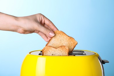 Woman taking roasted bread out of toaster on light blue background, closeup