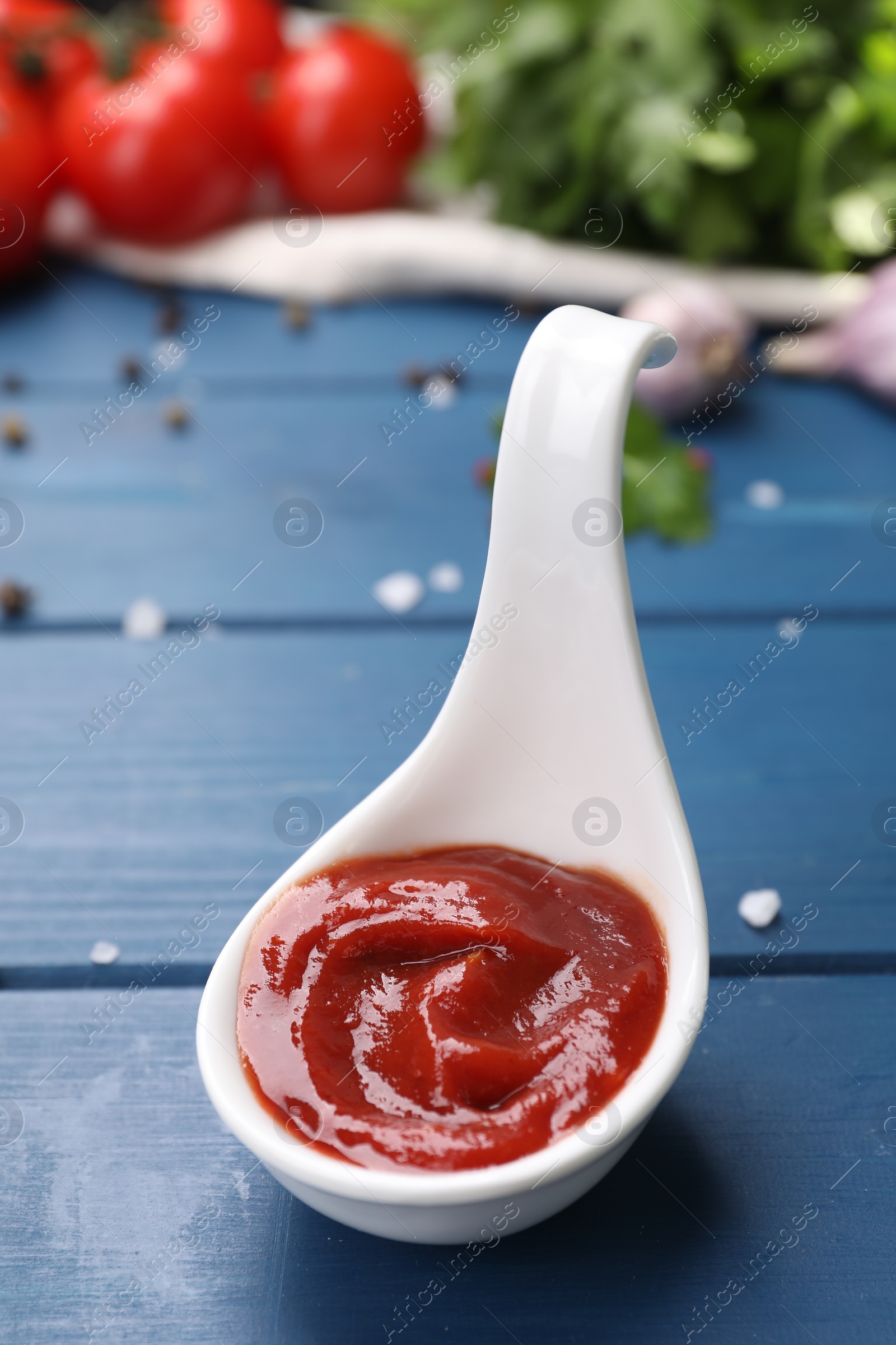 Photo of Organic ketchup in spoon and spices on blue wooden table, closeup. Tomato sauce