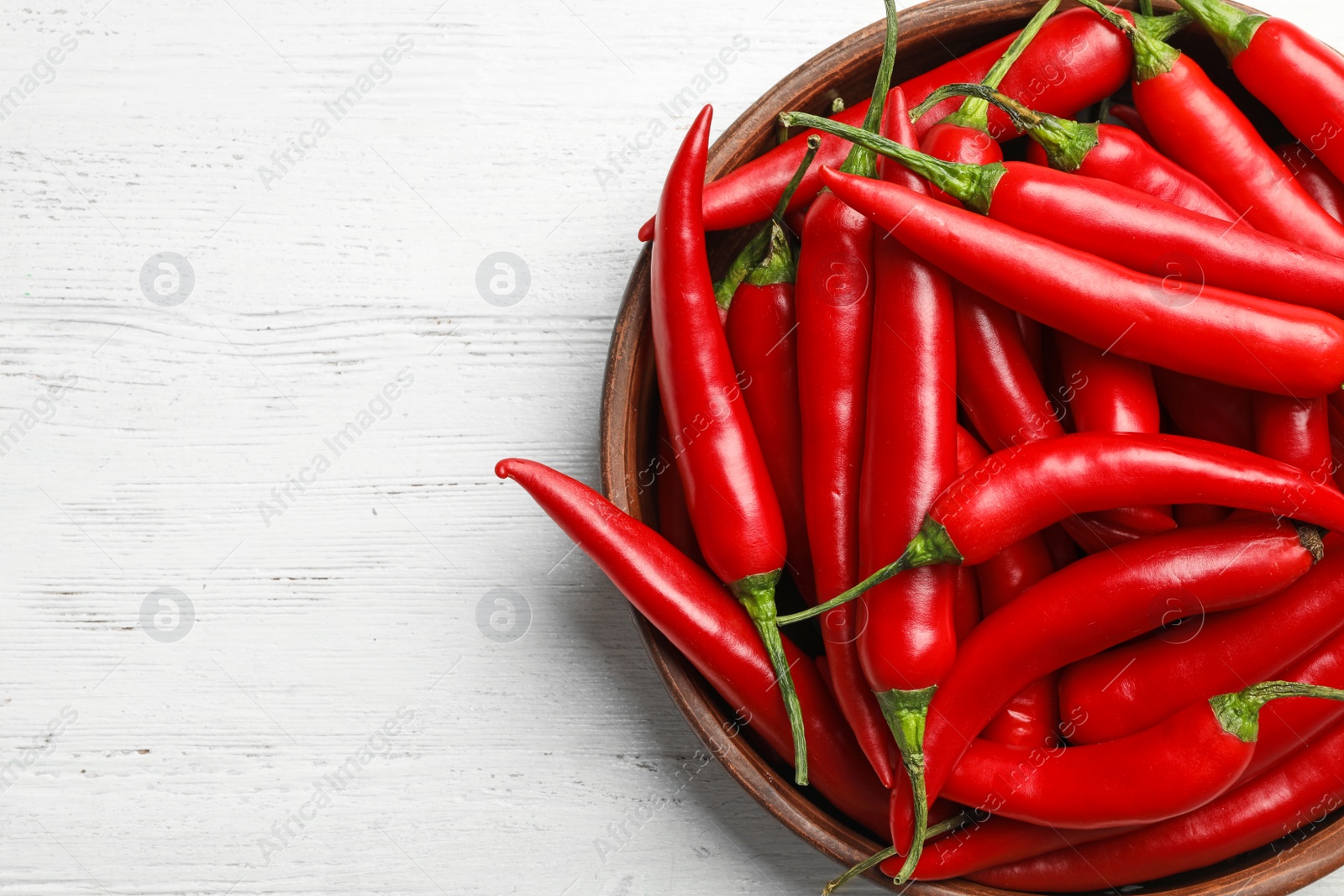 Photo of Wooden bowl with red hot chili peppers on white table, top view. Space for text