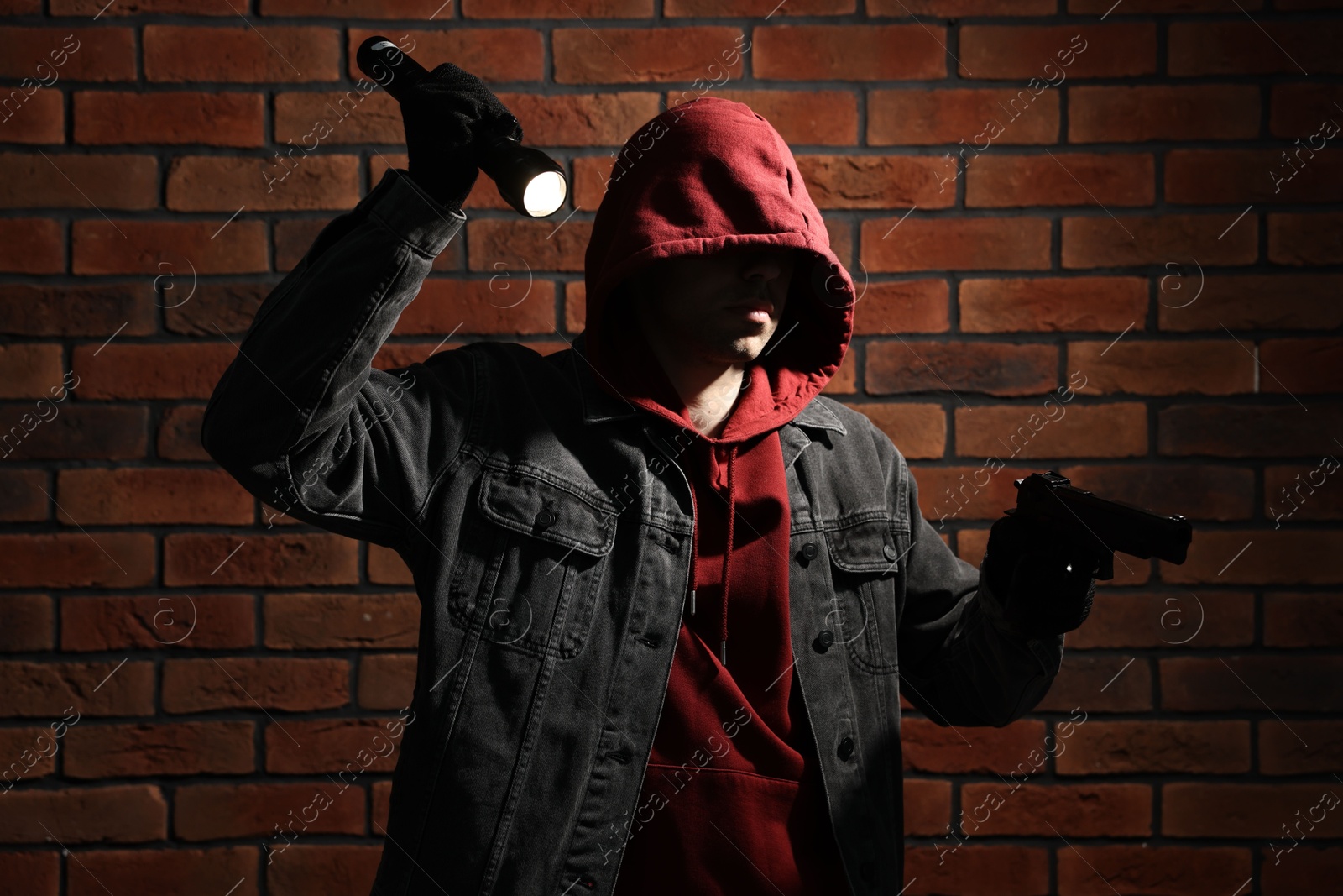 Photo of Thief in hoodie with gun and flashlight against red brick wall