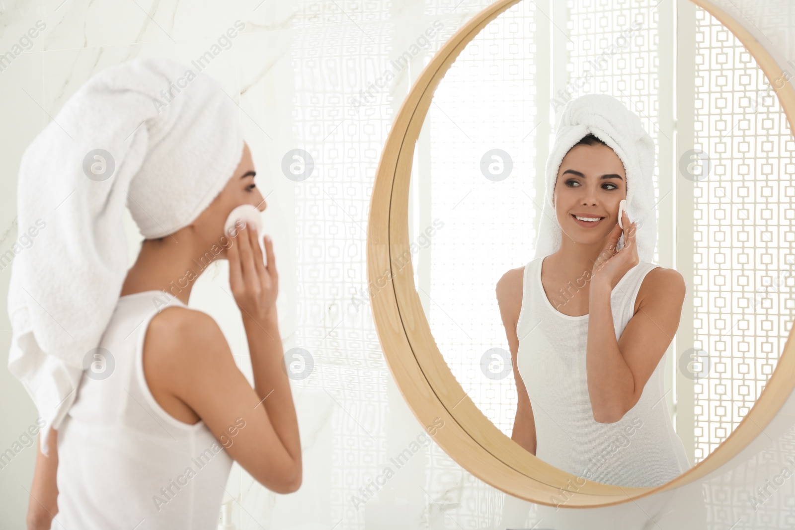 Photo of Happy young woman cleaning face with cotton pad near mirror in bathroom