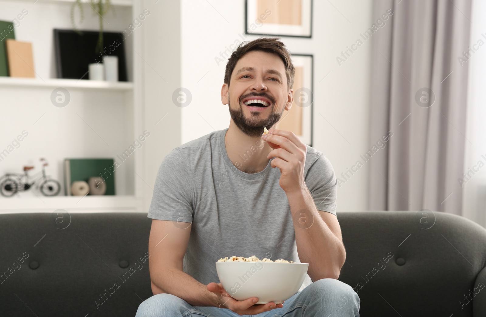 Photo of Happy man with bowl of popcorn watching movie via TV on sofa at home