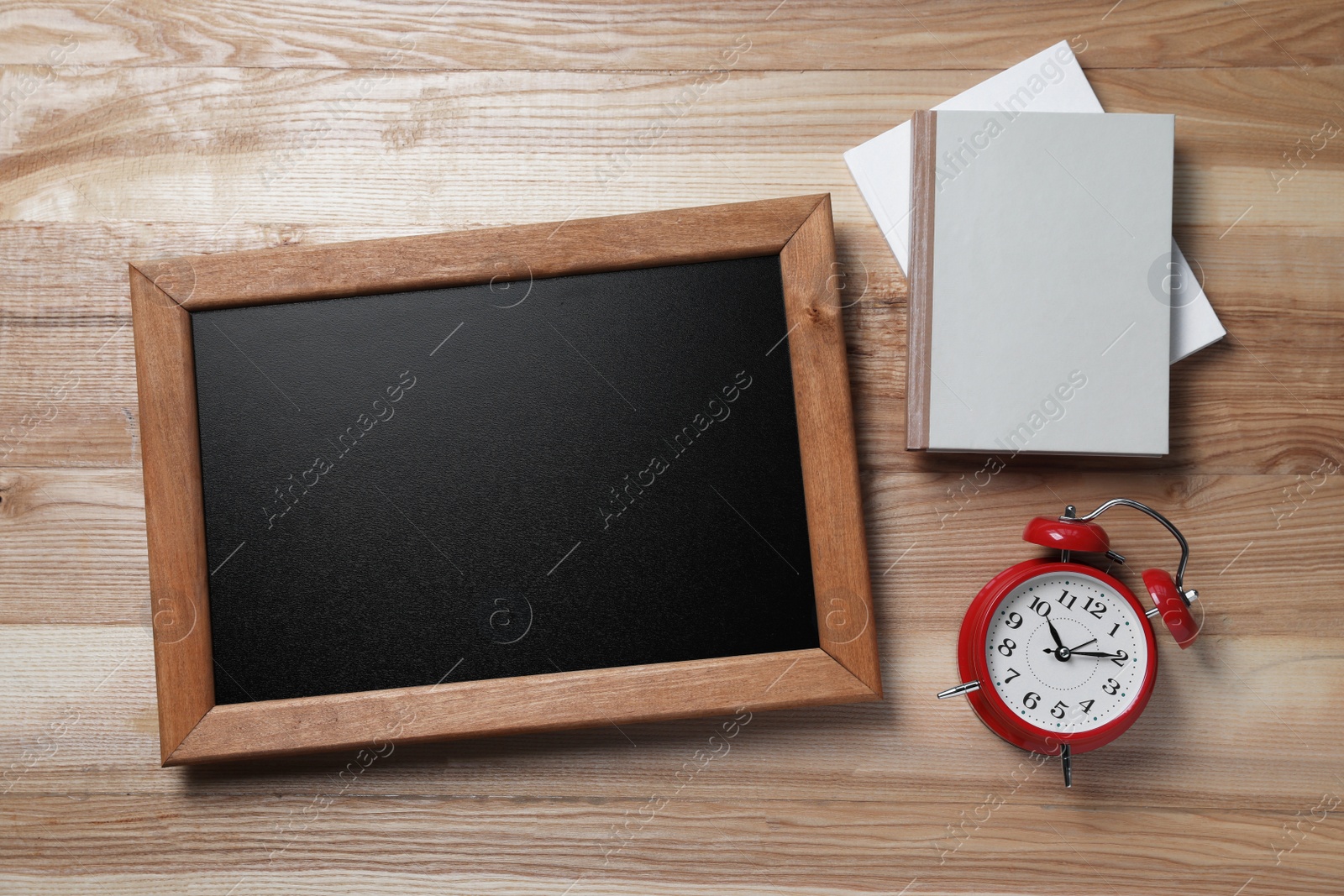 Photo of Blank chalkboard with notebooks and alarm clock on wooden table, flat lay. Space for text