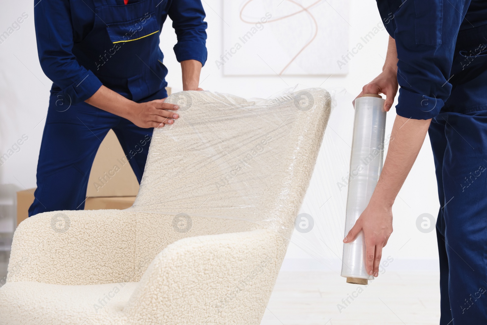 Photo of Male movers with stretch film wrapping armchair in house, closeup