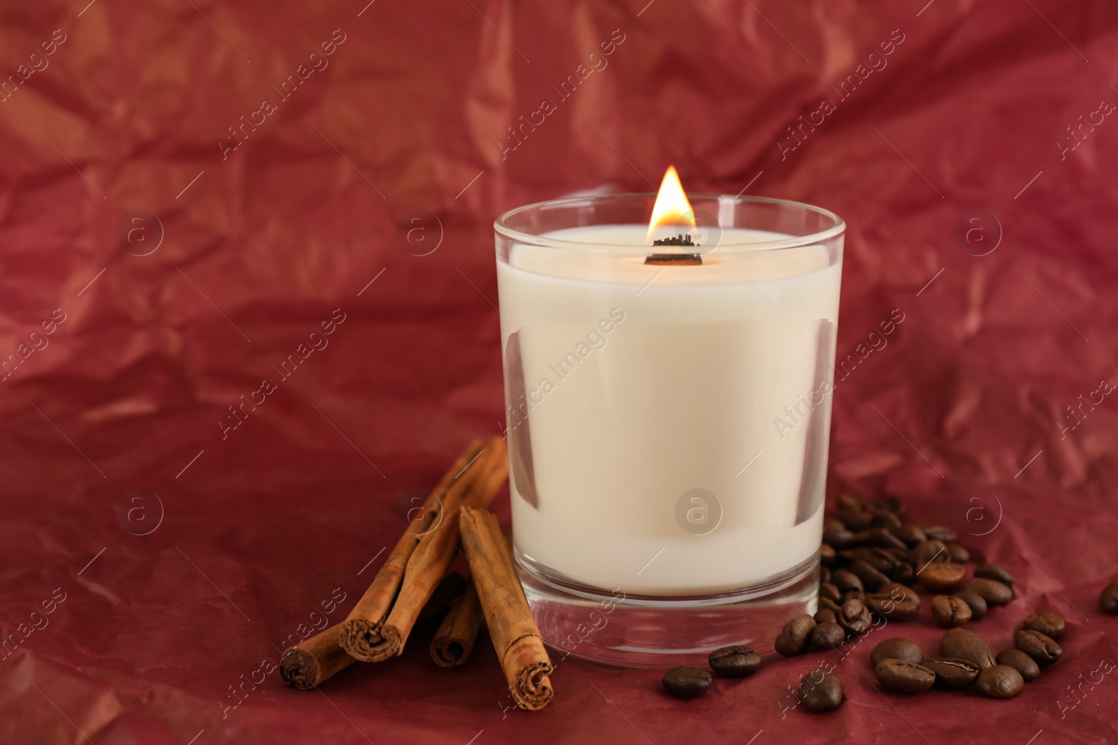 Photo of Burning soy candle, cinnamon sticks and coffee beans on crumpled red paper, space for text