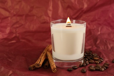 Burning soy candle, cinnamon sticks and coffee beans on crumpled red paper, space for text