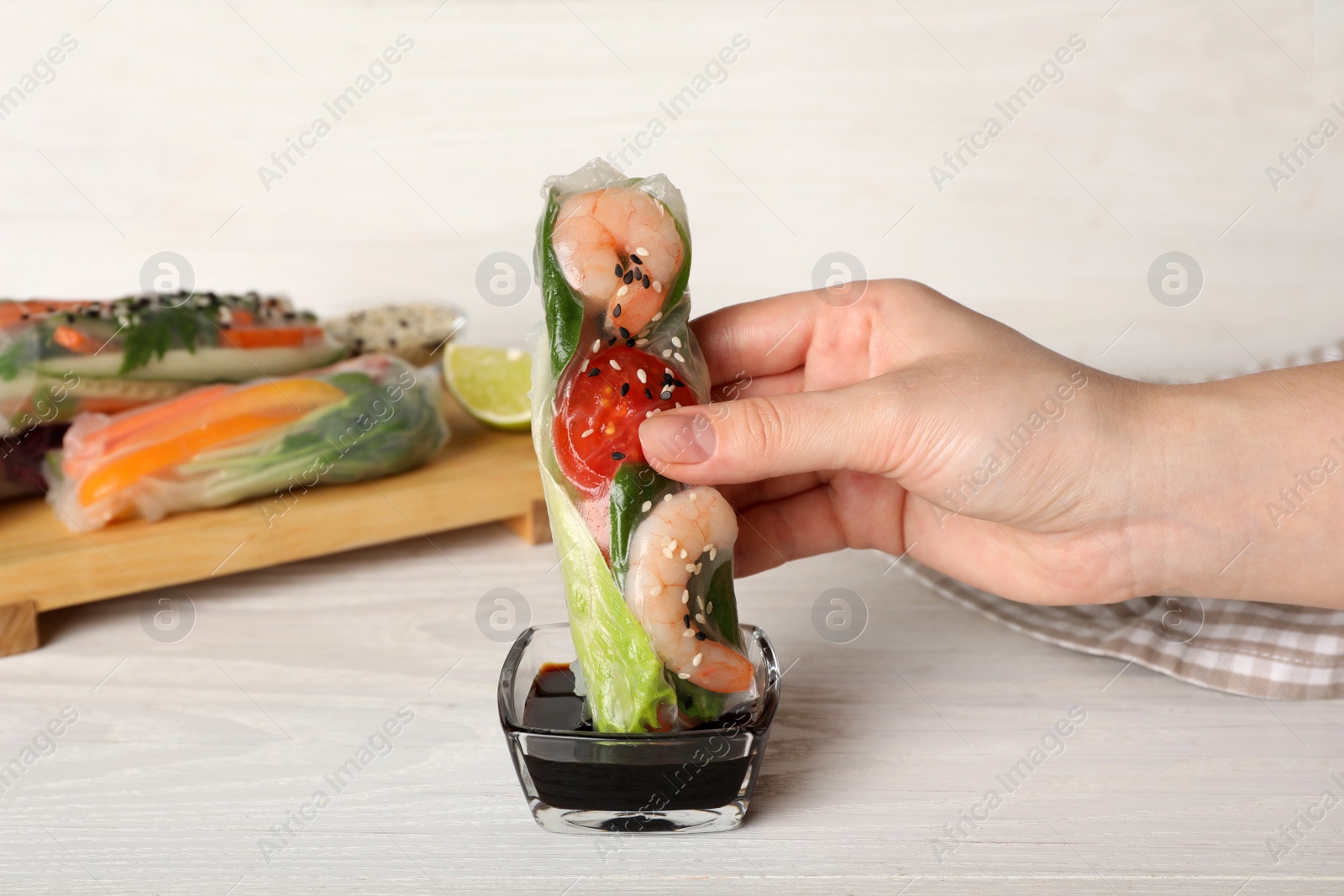 Photo of Woman dipping roll wrapped in rice paper into sauce at white wooden table, closeup