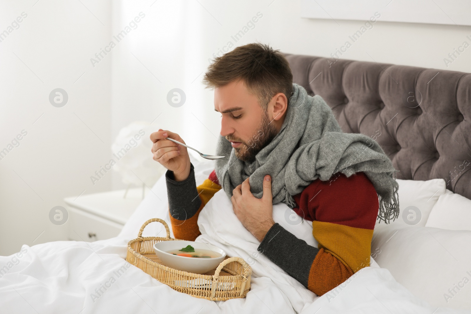 Photo of Sick young man with bowl of tasty soup in bed at home