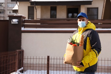 Courier in medical mask holding paper bag with groceries near house outdoors. Delivery service during quarantine due to Covid-19 outbreak