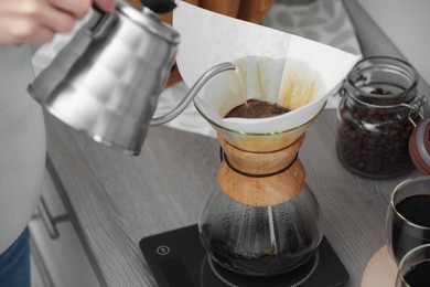 Photo of Woman pouring hot water into glass chemex coffeemaker with paper filter and coffee at countertop in kitchen, closeup