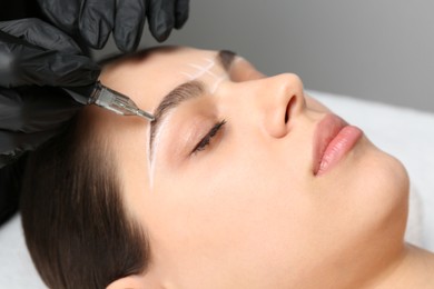 Photo of Beautician making permanent eyebrow makeup to young woman on grey background, closeup