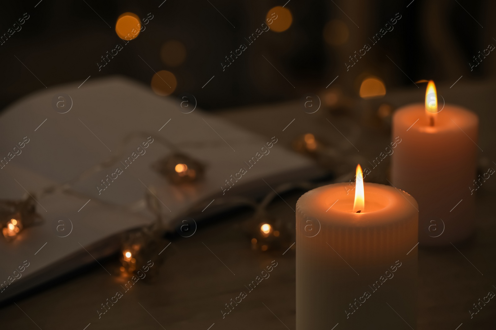 Photo of Burning candles, festive lights and open book in darkness, closeup. Space for text