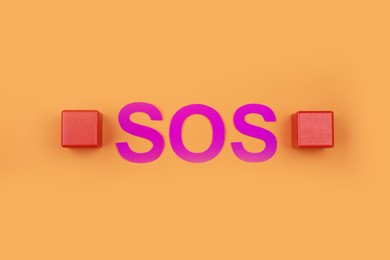 Photo of Abbreviation SOS made of paper letters and wooden cubes on orange background, flat lay