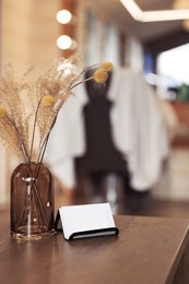 Photo of Barber business cards near glass vase with dried flowers on wooden table in hairdressing salon. Space for text