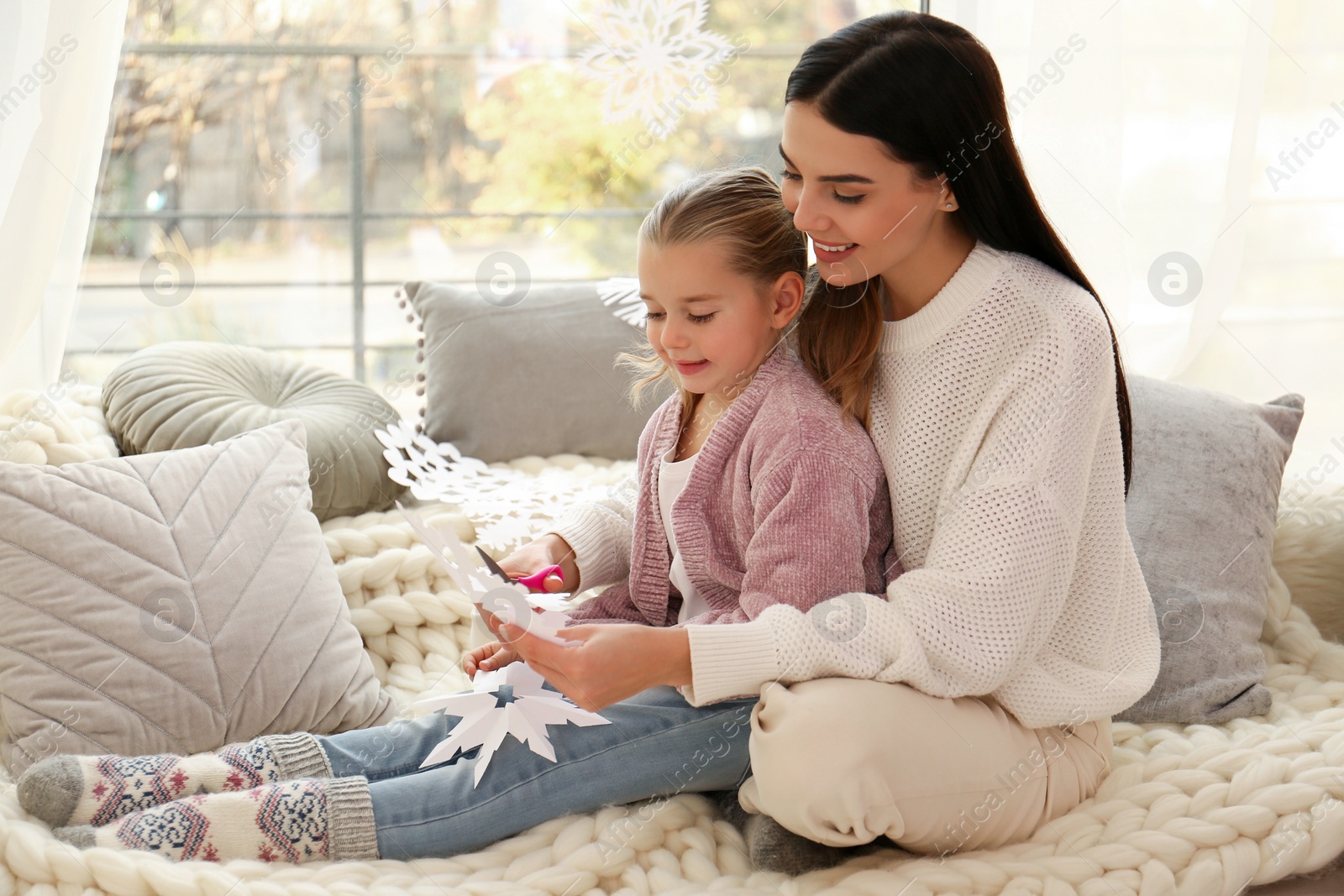 Photo of Mother and daughter making paper snowflakes near window at home