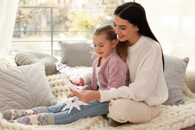 Photo of Mother and daughter making paper snowflakes near window at home