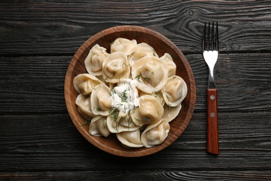 Photo of Tasty dumplings with sour cream in bowl on black wooden table, flat lay