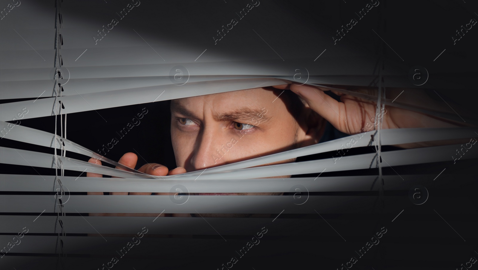 Image of Worried man looking through window blinds into darkness. Paranoia concept