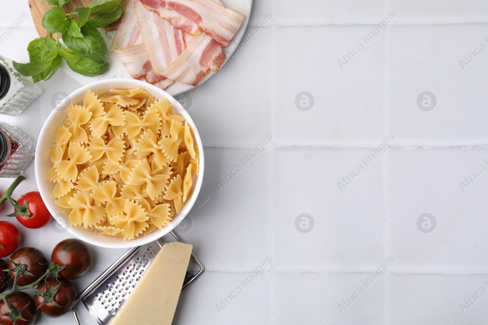 Photo of Raw pasta, bacon and fresh ingredients on white tiled table, flat lay. Space for text