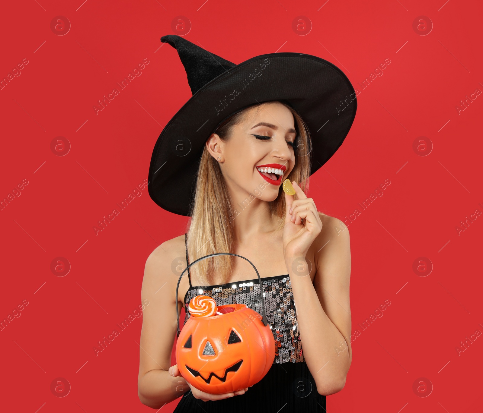 Photo of Beautiful woman wearing witch costume with Jack O'Lantern candy container on red background, space for text. Halloween party