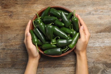 Young woman holding bowl with green hot chili peppers at wooden table, top view