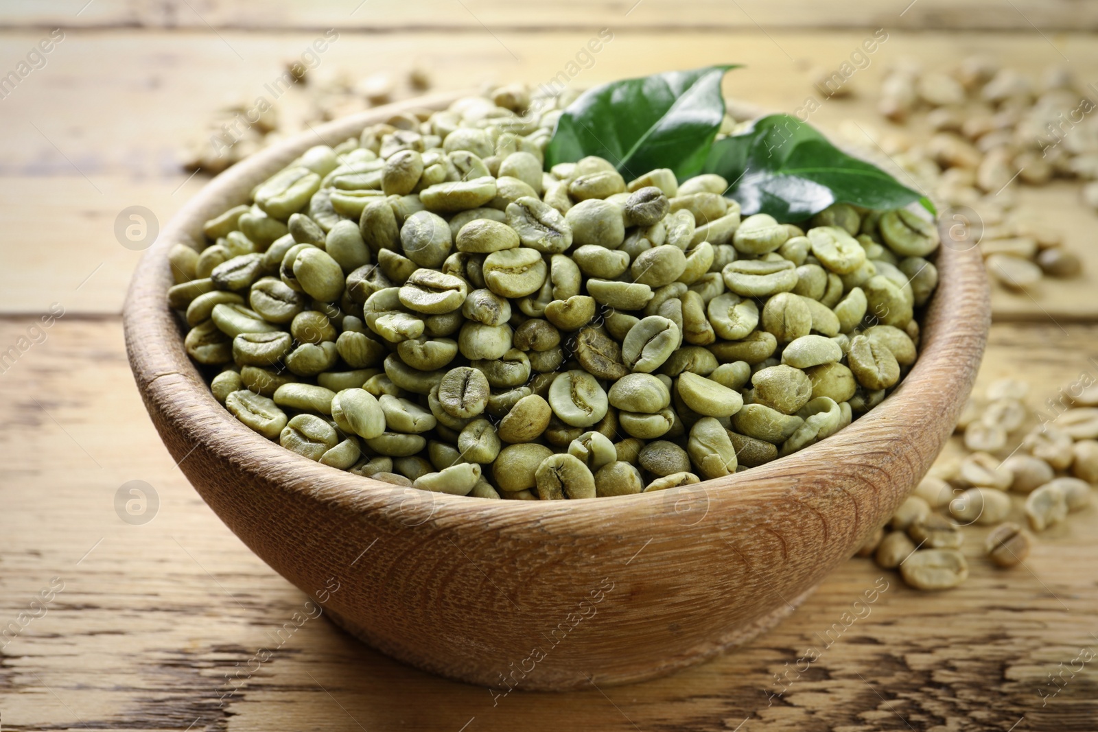 Photo of Green coffee beans and leaves in bowl on wooden table, closeup