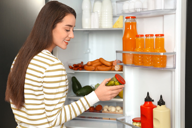 Photo of Young woman taking pickles out of refrigerator