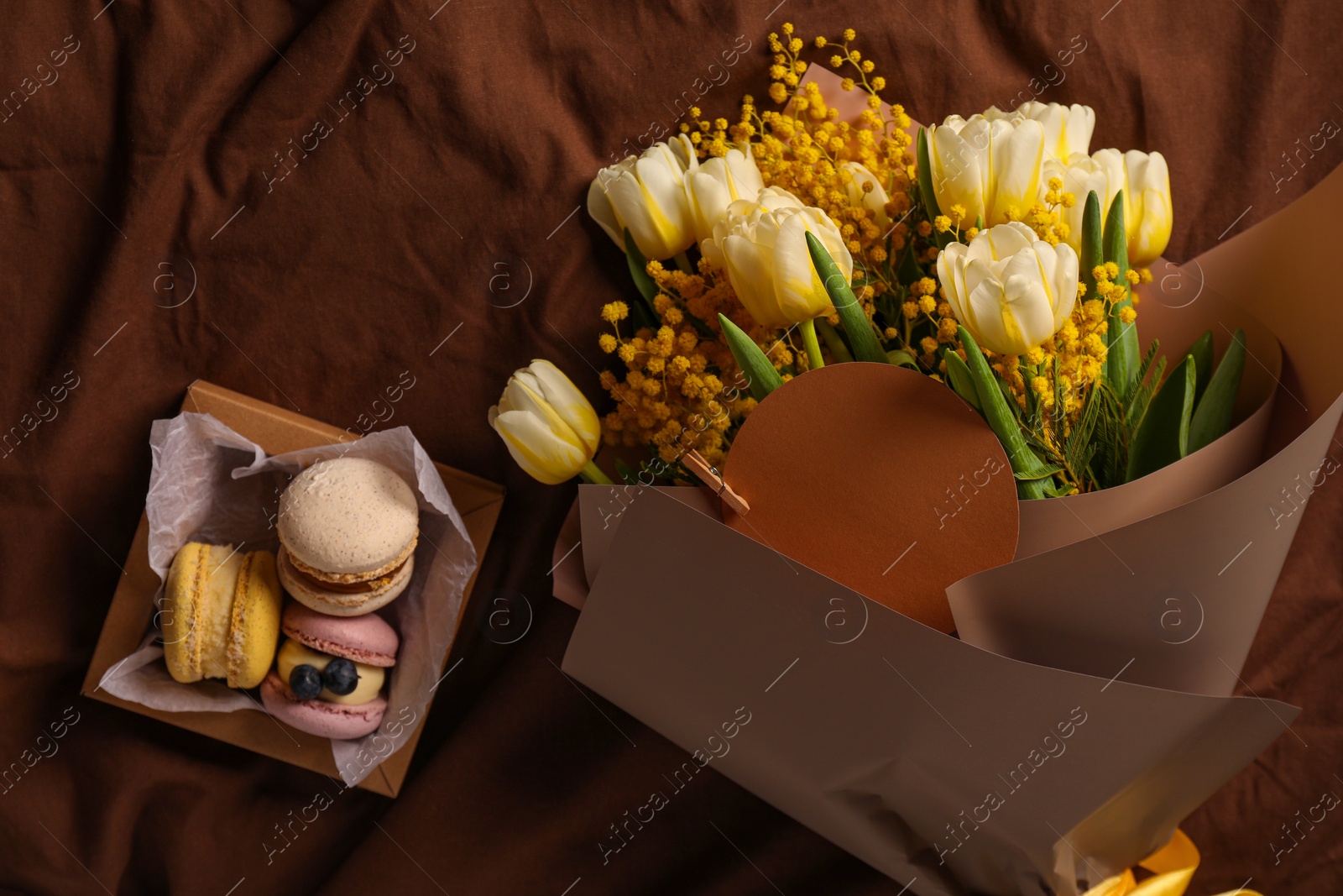 Photo of Bouquet of beautiful spring flowers, blank card and macarons on brown fabric, flat lay