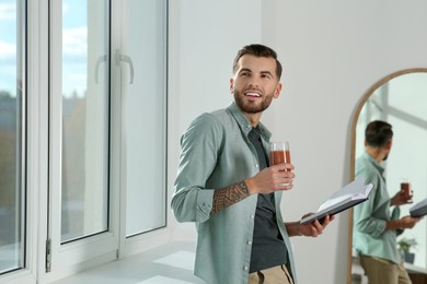 Photo of Handsome man with delicious smoothie and book near window indoors