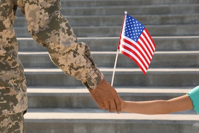 Photo of Soldier and his little daughter with American flag holding hands outdoors, closeup. Veterans Day in USA
