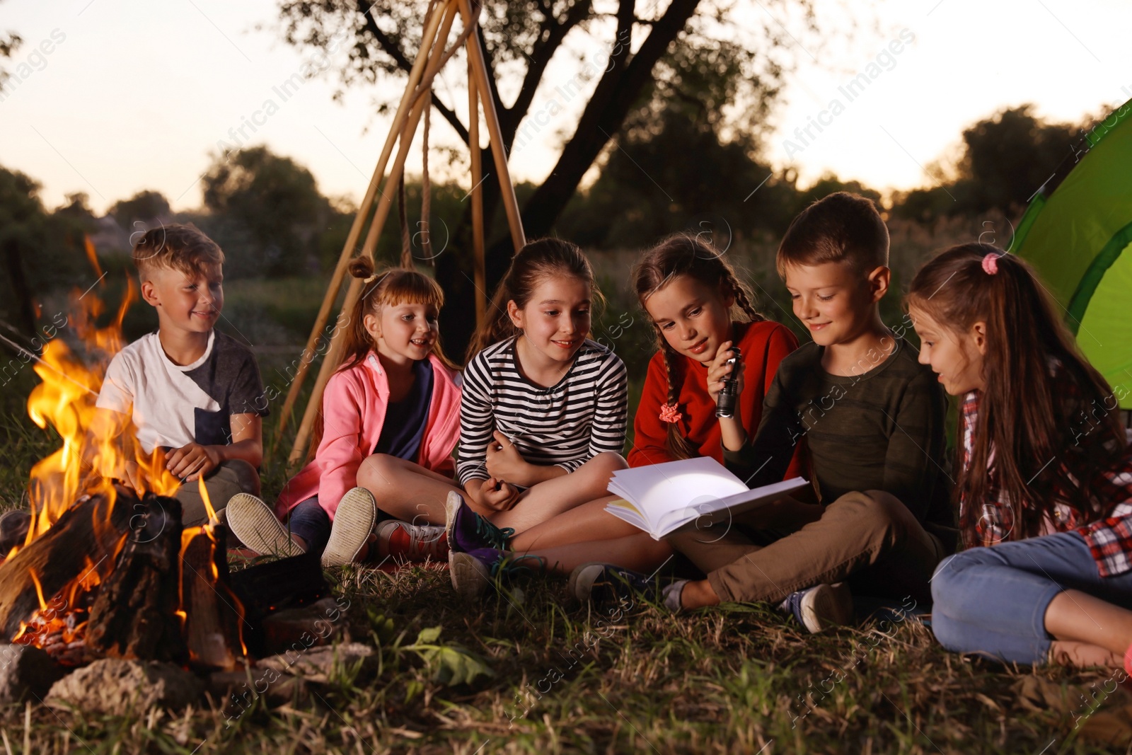 Photo of Little children reading book with flashlight outdoors. Summer camp