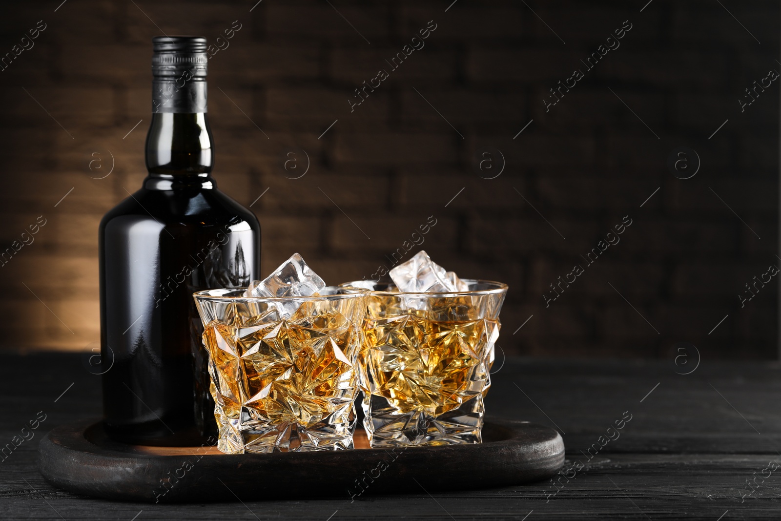 Photo of Whiskey with ice cubes in glasses and bottle on black wooden table, space for text