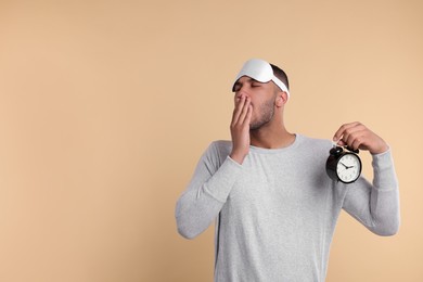Photo of Tired man with sleep mask and alarm clock yawning on beige background, space for text. Insomnia problem