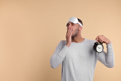 Tired man with sleep mask and alarm clock yawning on beige background, space for text. Insomnia problem