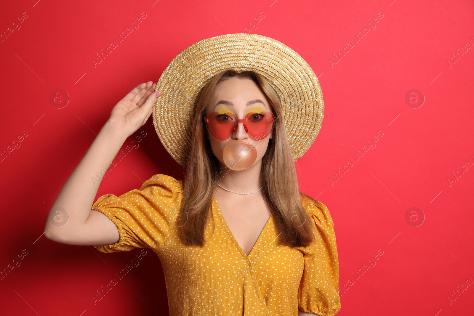 Photo of Fashionable young woman with bright makeup blowing bubblegum on red background