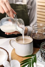 Woman pouring tasty coconut milk into mug of coffee at white table indoors, closeup