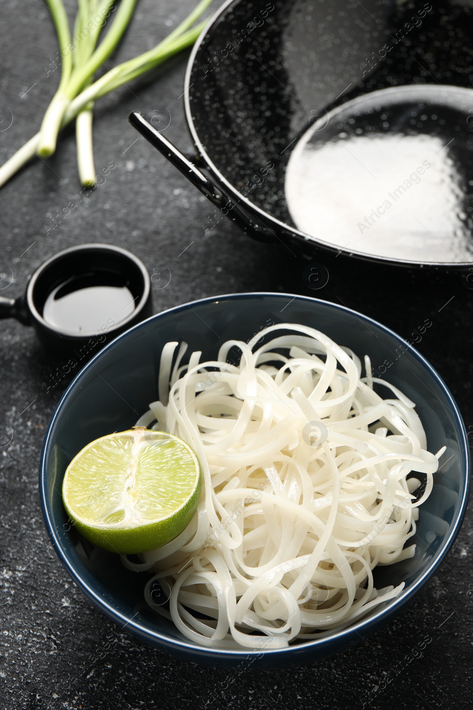 Photo of Noodles, lime and black wok on grey textured table, closeup
