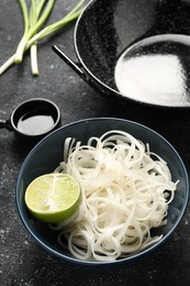 Photo of Noodles, lime and black wok on grey textured table, closeup