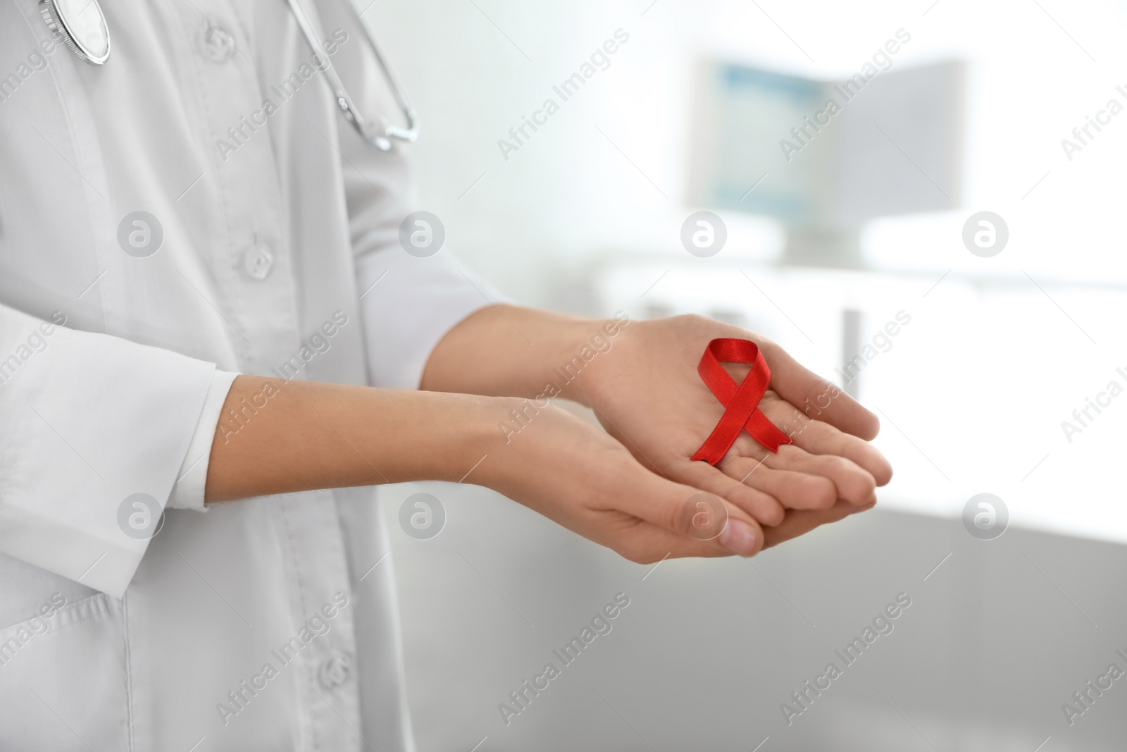 Photo of Doctor holding red ribbon on blurred background, closeup. AIDS awareness month