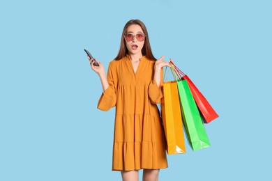 Photo of Surprised young woman with shopping bags and smartphone on light blue background. Big sale